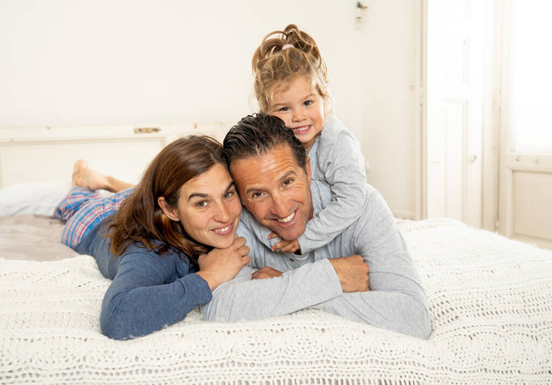 Портрет жизни прекрасной счастливой семьи, расслабляющей и проводящей время вместе в постели дома. Мама, папа, и милая дочка малыша весело целуются обнимаются и сближаются по утрам
. - Фото, изображение