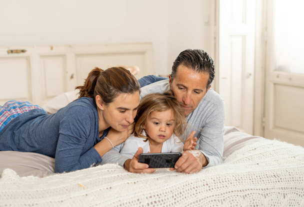 Mutlu bir aile evdeki yatakta cep telefonuyla internet üzerinden video izliyor. Anne, baba ve küçük kız akıllı telefonda oynuyorlar ve birlikte eğleniyorlar. Eğitim, dijital nesil ve teknolojide. - Fotoğraf, Görsel