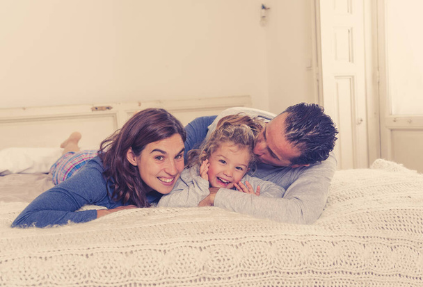 Lifestyle portrait of beautiful happy family relaxing and spending time together in bed at home. Mother, father, and cute toddler daughter having fun kissing hugging and bonding in the morning. - Foto, Imagem