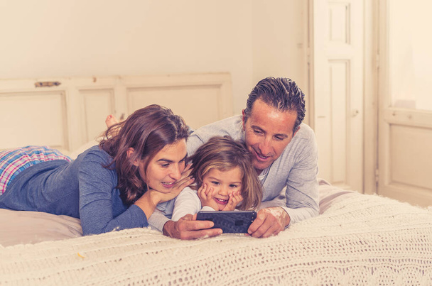 Ευτυχής οικογένεια βλέποντας βίντεο στο διαδίκτυο στο κινητό τηλέφωνο στο κρεβάτι στο σπίτι. Η μαμά μπαμπάς και το κοριτσάκι παίζουν στο smartphone διασκεδάζοντας μαζί. Στην Εκπαίδευση, την ψηφιακή παραγωγή και την τεχνολογία. - Φωτογραφία, εικόνα