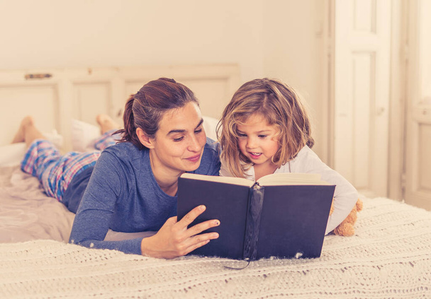 Young Mother and cute little daughter reading a book together. Lifestyle image of happy mom and cute blonde girl having fun reading fairy tales in bed. In happy family, parenting and education concept. - Fotó, kép