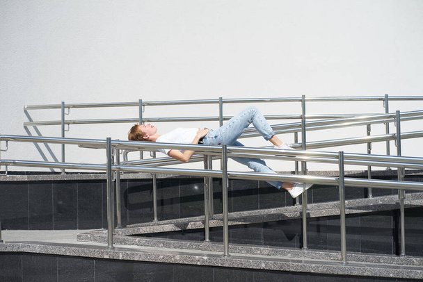 Shorthaired redhead skinny caucasian young woman lies on railing in urban center in hot summer day - 写真・画像