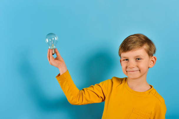 Il ragazzo tiene in mano una lampadina e sorride, un bambino in abiti gialli su sfondo blu
. - Foto, immagini
