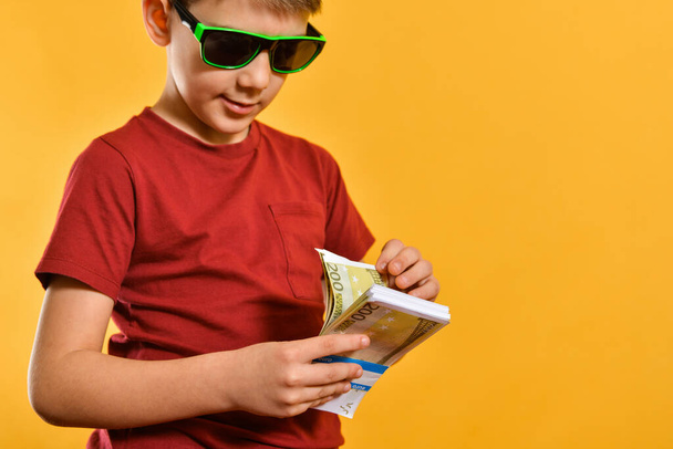 Ein Junge mit Sonnenbrille betrachtet Banknoten im Bündel, ein glamouröses und stilvolles Kind mit Währung auf gelbem Hintergrund. - Foto, Bild
