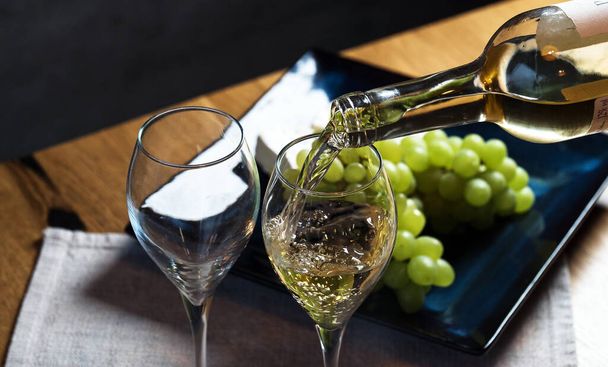 Pouring white wine into the glasses above wooden table background, set with grape and cheese  - Fotoğraf, Görsel