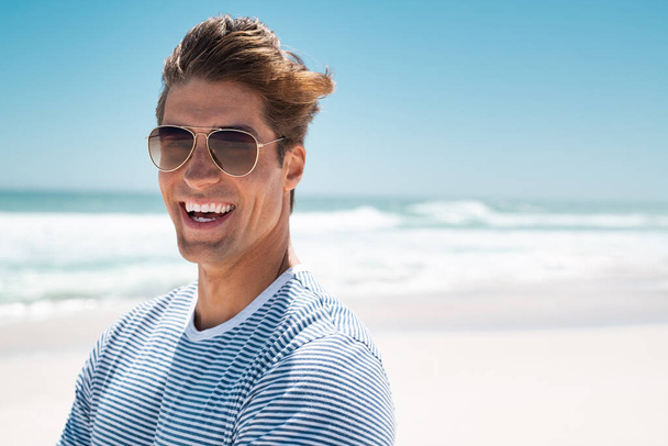 Portrait of handsome young man wearing sunglasses and looking at camera. Portrait of joyful man enjoying summer holiday at beach. Young casual man in shades with big grin and copy space. - Zdjęcie, obraz