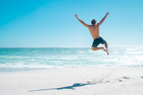 Back view of happy young man jumping with outstretched arms on the beach with copy space. Rear view of shirtless guy wearing swimwear and having fun on tropical beach during vacation. Carefree and freedom concept. - Foto, Imagem