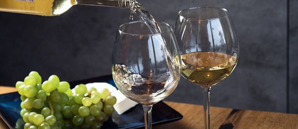 Fehér bor öntése a poharakba a fából készült asztal háttere felett, szőlővel és sajttal díszítve  - Fotó, kép