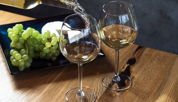 Pouring white wine into the glasses above wooden table background, set with grape and cheese  - Photo, Image