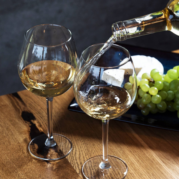 Gießen Weißwein in die Gläser über hölzernem Tischhintergrund, mit Trauben und Käse  - Foto, Bild