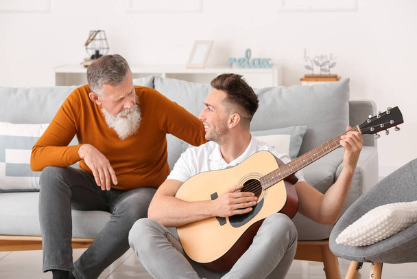 Ο μεγαλύτερος άντρας και ο ενήλικος γιος του παίζουν κιθάρα στο σπίτι. - Φωτογραφία, εικόνα