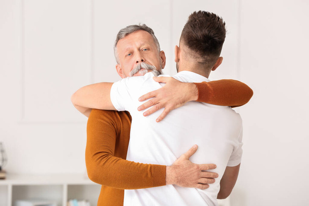 Ο μεγαλύτερος άντρας και ο ενήλικος γιος του αγκαλιάζονται στο σπίτι. - Φωτογραφία, εικόνα