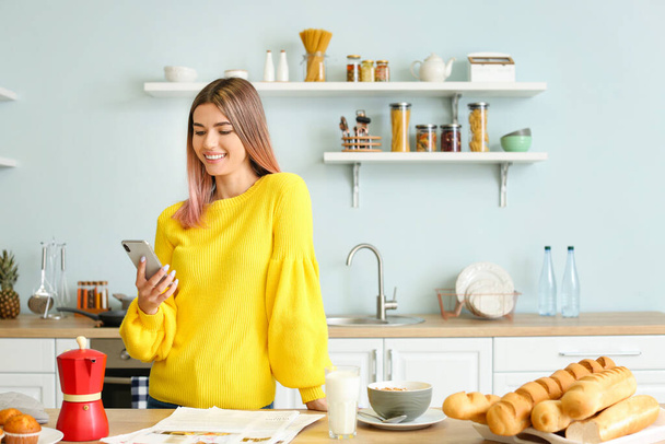 Beautiful young woman with mobile phone in kitchen - 写真・画像