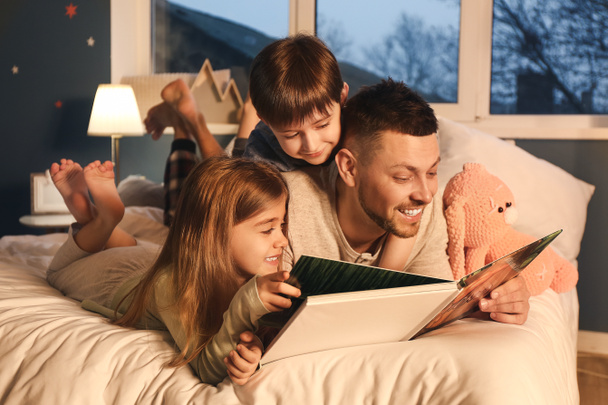 Isä ja hänen pienet lapsensa lukevat iltasatua kotona.
 - Valokuva, kuva