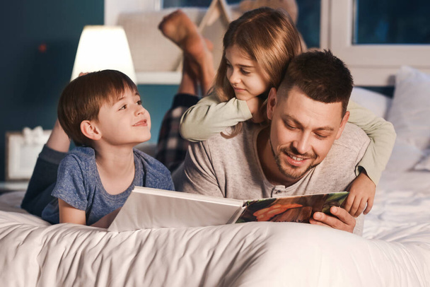 Padre y sus pequeños hijos leyendo cuentos para dormir en casa
 - Foto, Imagen