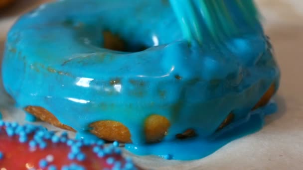 Uma fileira de rosquinhas fritas redondas maiores em uma mesa de culinária caseira. Uma escova de silicone de cozinha especial aplica um esmalte azul brilhante à superfície do donut. Gordo, junk food, fast food close-up
 - Filmagem, Vídeo