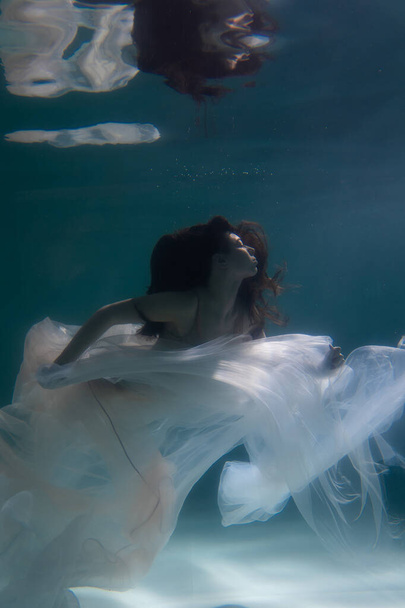Beautiful girl swims underwater with long hair. Blue or gold background like gold. The atmosphere of a fairy tale or magic. Diving under the water with a shiny cloth - Foto, afbeelding