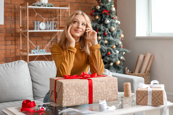 Beautiful young woman talking by phone while wrapping Christmas gifts at home - Photo, Image
