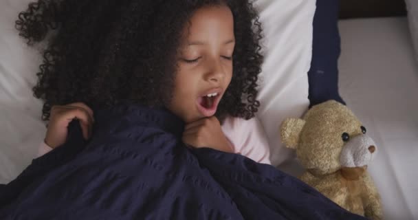High angle front view of a mixed race girl enjoying time at home, lying in bed, yawning, covering herself with a duvet, in slow motion - Materiaali, video