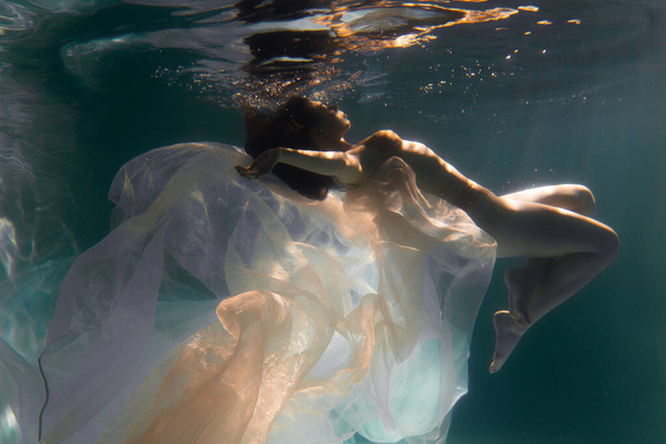 Beautiful girl swims underwater with long hair. Blue or gold background like gold. The atmosphere of a fairy tale or magic. Diving under the water with a shiny cloth - Foto, afbeelding