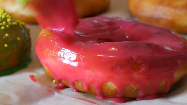 A row of larger round freshly fried donuts on a home cooking table. A special kitchen silicone brush applies a bright pink glaze to the surface of the donut. Fatty, junk food, fast food - Footage, Video