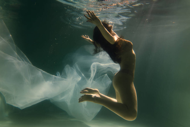 Piękna dziewczyna pływa pod wodą z długimi włosami. Niebieskie lub złote tło jak złoto. Atmosfera bajki lub magii. Nurkowanie pod wodą z błyszczącą szmatką - Zdjęcie, obraz