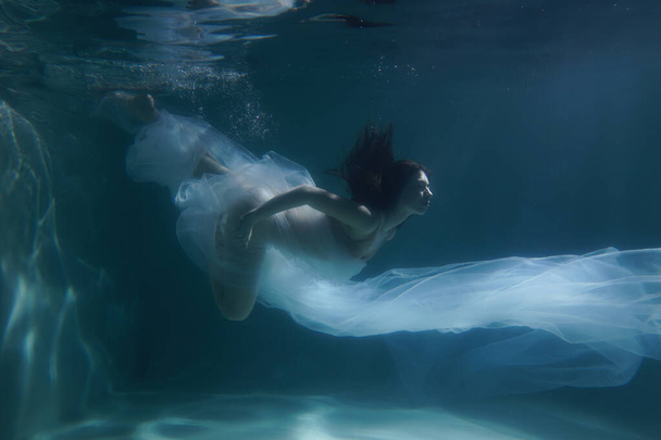 Beautiful girl swims underwater with long hair. Blue or gold background like gold. The atmosphere of a fairy tale or magic. Diving under the water with a shiny cloth - Fotó, kép