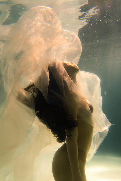 Beautiful girl swims underwater with long hair. Blue or gold background like gold. The atmosphere of a fairy tale or magic. Diving under the water with a shiny cloth - Foto, immagini