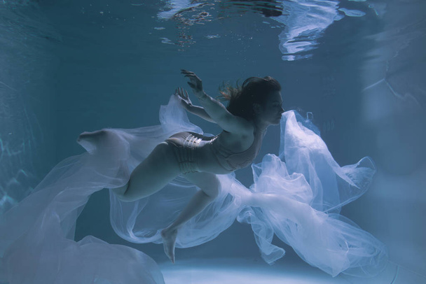 Beautiful girl gymnast and sportswoman swims underwater with shining fabric in the pool	 - Photo, Image