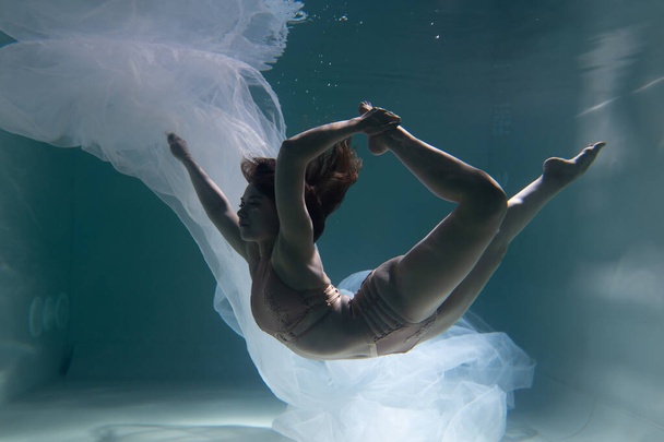 Beautiful girl gymnast and sportswoman swims underwater with shining fabric in the pool	 - 写真・画像