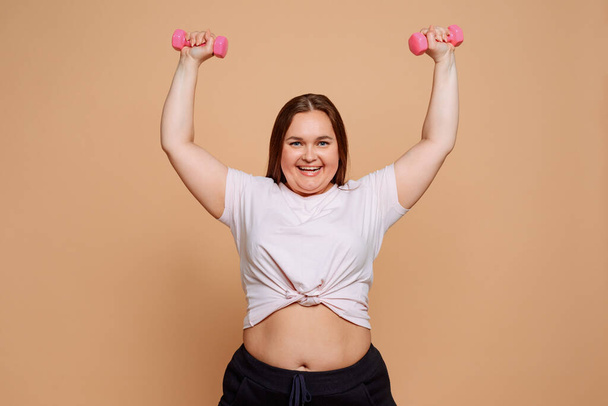 Delighted young overweight girl doing overhead dumbbell exercises - Photo, image