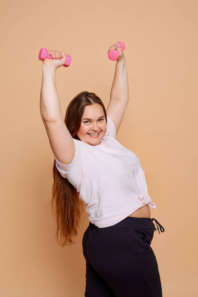Beautiful plus size model posing with pink dumbbells on beige background - Fotoğraf, Görsel