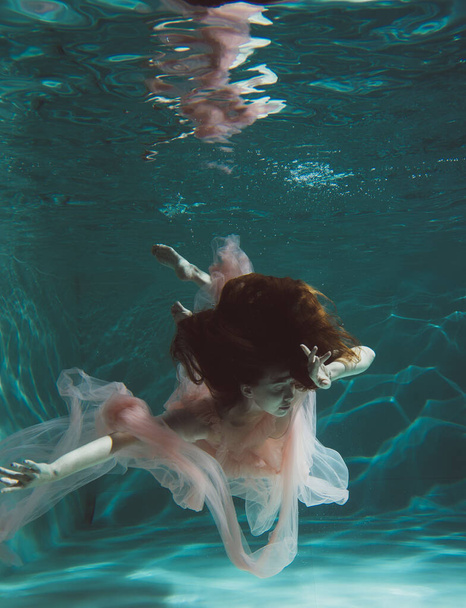 A girl with long dark hair swims underwater in a pink dress and with a crown on her head, like an underwater queen. Fairy tale suitable for advertising - Φωτογραφία, εικόνα