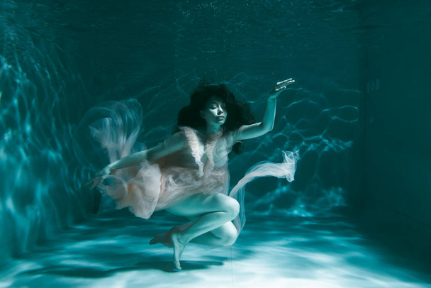 A girl with long dark hair swims underwater in a pink dress and with a crown on her head, like an underwater queen. Fairy tale suitable for advertising - Фото, изображение