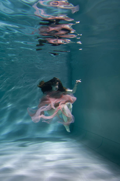 A girl with long dark hair swims underwater in a pink dress and with a crown on her head, like an underwater queen. Fairy tale suitable for advertising - Fotoğraf, Görsel