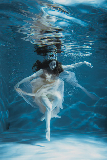 Une fille aux longs cheveux foncés nage sous l'eau dans une robe rose et avec une couronne sur la tête, comme une reine sous-marine. Conte de fées adapté à la publicité
 - Photo, image