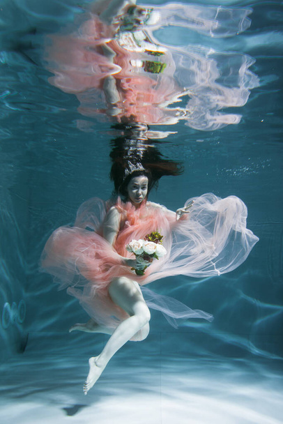A girl with long dark hair swims underwater in a pink dress and with a crown on her head, like an underwater queen. Fairy tale suitable for advertising - Foto, afbeelding