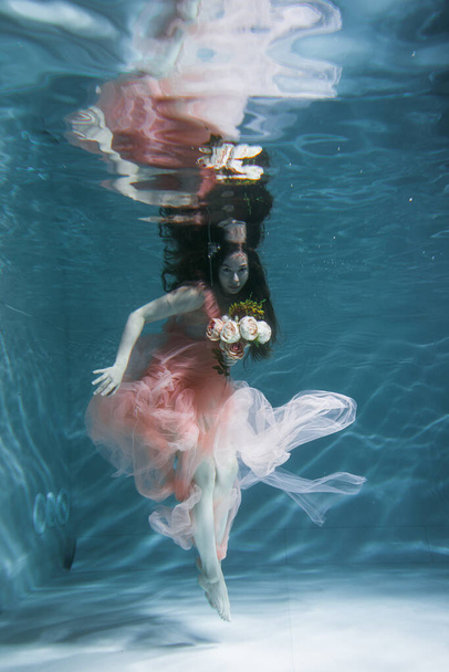 A girl with long dark hair swims underwater in a pink dress and with a crown on her head, like an underwater queen. Fairy tale suitable for advertising - Photo, image