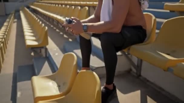 Egy vonzó, fiatal, félmeztelen sportoló törülközővel a vállán ül és vizet iszik a stadionban, félrenéz. - Felvétel, videó