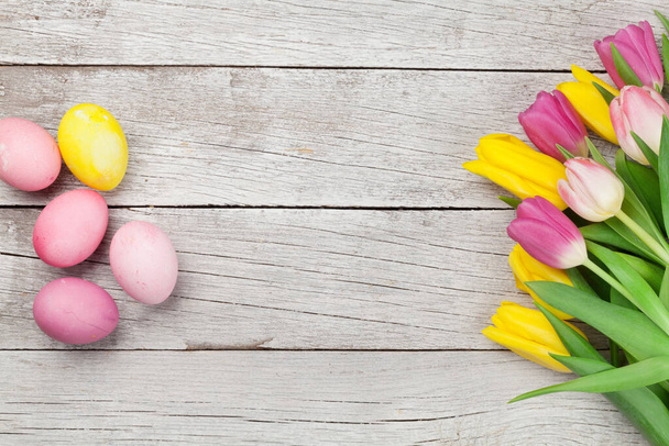 Easter greeting card with easter eggs and tulip flowers over wooden background. Top view flat lay with copy space - 写真・画像