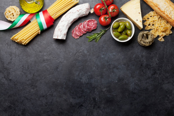 Italian cuisine food ingredients. Pasta, cheese, salami, olives and tomatoes. Top view flat lay on stone table with copy space - Photo, Image