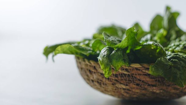 Conceito de comida vegan saudável: backlight close-up com efeito bokeh de folhas de espinafre verde recém-colhidas colocadas em uma cesta de vime em uma mesa de madeira branca e fundo branco
 - Foto, Imagem