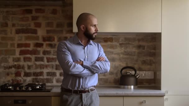  A bald brutal businessman with a beard folds his arms over his chest turns and looks straight. A man wears two silver earrings, a blue shirt, grey trousers, and a brown leather belt in the kitchen. - Záběry, video