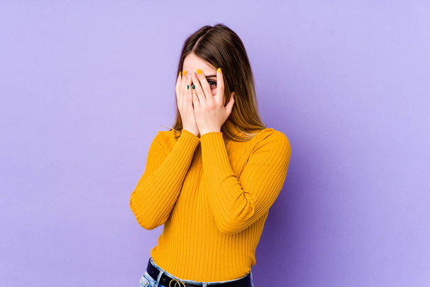 Mladá běloška izolovaná na purpurovém pozadí bliká na kameru skrz prsty, rozpačitá zakrývající tvář. - Fotografie, Obrázek