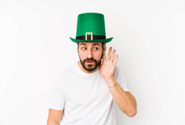 Young caucasian man wearing a saint patricks hat isolated trying to listening a gossip. - Photo, image