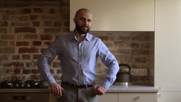 A bald brutal businessman with a beard and smile folds his arms over his chest in the kitchen. A man wears a blue shirt, grey trousers, and a brown leather belt. - Záběry, video