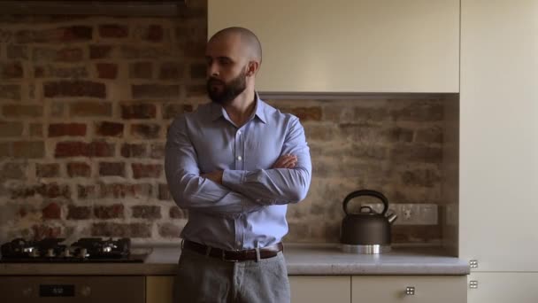 A bald brutal businessman with a beard folds his arms over his chest turns his head into left. A man wears two silver earrings, a blue shirt, grey trousers, and a brown leather belt in the kitchen. - Filmmaterial, Video