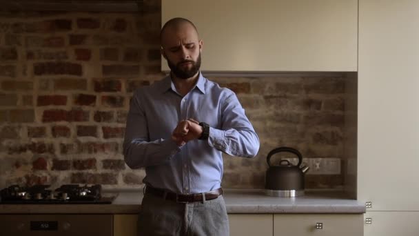 A bald brutal businessman with a beard in a blue shirt, grey trousers looks on his wristwatch, then puts his hands in his pockets in the kitchen - 映像、動画