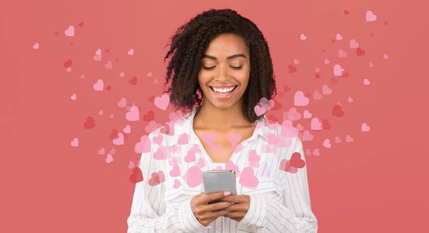 Donna afroamericana browsign attraverso i social media sul telefono cellulare con cuori che volano intorno, sfondo rosa
 - Foto, immagini