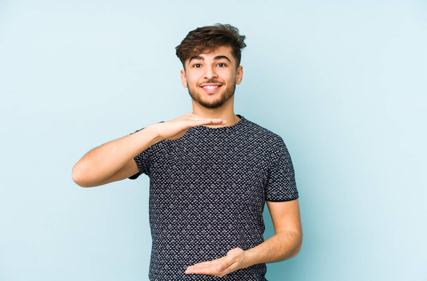 Jeune homme arabe isolé sur un fond bleu tenant quelque chose avec les deux mains, présentation du produit
. - Photo, image